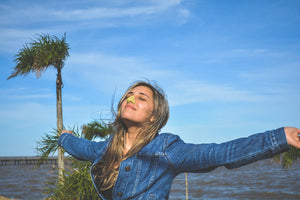 Girl outside smiling wearing denim jacket in front of blue sky and body of water with yellow NÖZ brand sunscreen on her nose